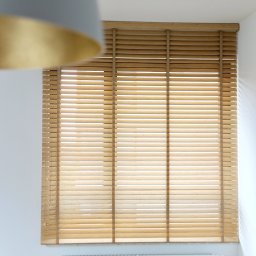 Mimo Decor - Jasne, drewniane osłony okienne z poziomymi lamelami, zamontowane w białej wnęce nad kaloryferem. W tle rozmyta szaro-złota lampa.