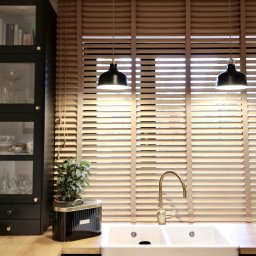 Mimo Decor - Elegancka kuchnia z oknem zasłoniętym poziomymi, drewnianymi lamelami, białym podwójnym zlewem i złotą, wygiętą baterią. Widoczne czarne szafki i drewniany blat.