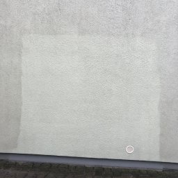 Czyścioch AK - Fragment jasnej ściany z fakturą, gdzie centralny obszar jest wyraźnie jaśniejszy i czystszy niż boczne, z białą kratką wentylacyjną u dołu i mokrą kostką brukową.
