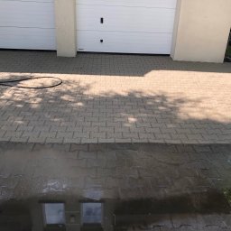 Czyścioch AK - Kostka brukowa przed dwoma białymi bramami garażowymi, częściowo wyczyszczona, z wyraźną linią podziału między mokrą a suchą nawierzchnią. W tle widoczne narzędzie do czyszczenia.