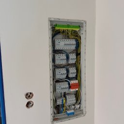 M.S Elektro - Mikołaj Smolik - Otwarta rozdzielnica elektryczna w białej ścianie, z rzędami wyłączników, pomarańczowym ogranicznikiem przepięć i uporządkowanym okablowaniem. Obok dwa otwory na gniazdka.