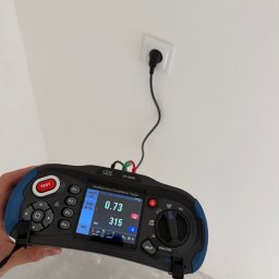 M.S Elektro - Mikołaj Smolik - Ręka trzyma wielofunkcyjny tester instalacji CEM DT-6650, pokazujący na ekranie wartości 0.73 i 315, podłączony do białego gniazdka ściennego.