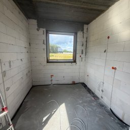 M.S Elektro - Mikołaj Smolik - Niewykończone pomieszczenie z białymi ścianami z bloczków, betonowym sufitem i podłogą. Widoczne okno. Na ścianach poprowadzone peszle i puszki pod okablowanie. Na podłodze zwinięty biały przewód.