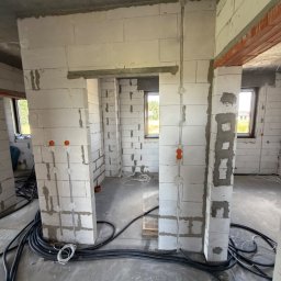 M.S Elektro - Mikołaj Smolik - Niezatynkowane wnętrze budynku z białych bloczków, z widocznymi rurkami instalacji elektrycznej, pomarańczowymi puszkami i kablami na betonowej posadzce. W tle okna z zielenią.