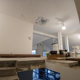 Wykonczenia wnetrz - Remontowane wnętrze z białymi ścianami i sufitem, widoczne wiadra gładzi Knauf Super Finish, betonowa antresola i odsłonięte przewody oświetleniowe.