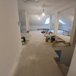Wykonczenia wnetrz - Poddasze w trakcie prac wykończeniowych, z gładkimi, białymi ścianami i oknami dachowymi. Na betonowej posadzce wiadra Knauf, materiały budowlane i tymczasowe oświetlenie.