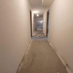 Wykonczenia wnetrz - Wąski korytarz w trakcie prac wykończeniowych, z idealnie gładkimi, jasnymi ścianami, surową betonową podłogą i widocznymi drewnianymi ościeżnicami.
