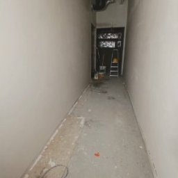 Wykonczenia wnetrz - Wąski korytarz z jasnymi, równymi ścianami po przygotowaniu, widocznymi rurami pod sufitem, drabiną i wiadrami na końcu.