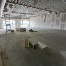 Wykonczenia wnetrz - Duże, puste pomieszczenie biurowe w trakcie remontu, ze ścianami z płyt gipsowo-kartonowych po szpachlowaniu, widocznym rusztowaniem i workami z zaprawą. Odsłonięty sufit z rurami wentylacyjnymi.