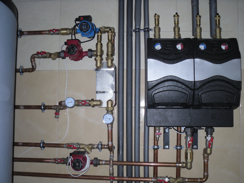 Kompleksowa instalacja kotłowni z dwoma rozdzielaczami, pompami obiegowymi i manometrami, widoczne rury miedziane i stalowe, zamontowane na ścianie wyłożonej płytkami.