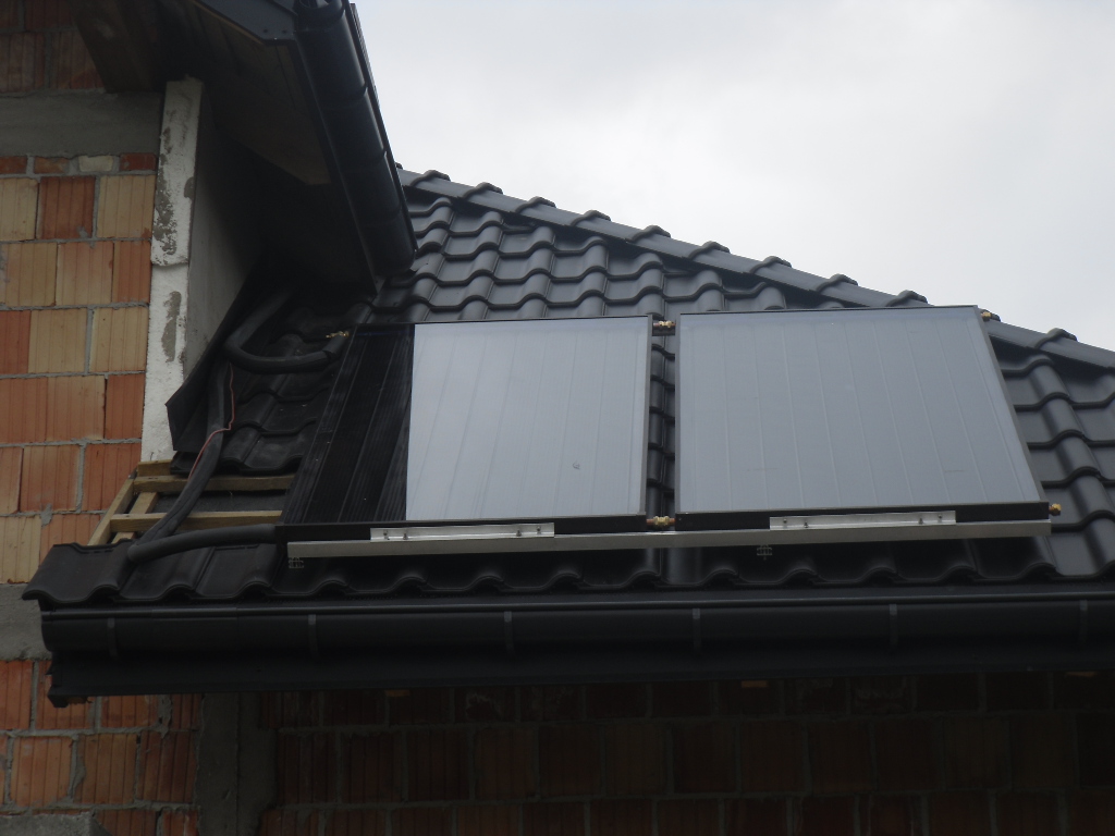 Dwa panele solarne zamontowane na czarnym dachu z dachówki ceramicznej, widoczne rury doprowadzające do paneli, mur z cegły dziurawki w tle.