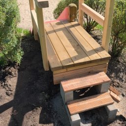 Lucjan Buduje - Nowo zbudowany drewniany mostek z poręczami i dwoma stopniami, prowadzący na piaszczysty plac zabaw. Świeże drewno, widoczne śruby, ziemia wokół.