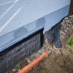 Lucjan Buduje - Szara ściana fundamentowa z tynkiem, czarną izolacją kubełkową i pomarańczową rurą drenażową w wykopie. Widoczne kamienie i ukośne pasma światła słonecznego na elewacji.