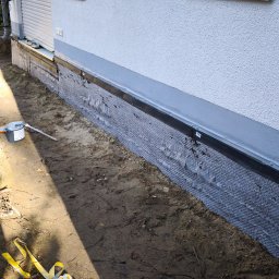 Lucjan Buduje - Izolacja fundamentów budynku z widoczną folią kubełkową i częściowo zasypanym wykopem. Jasna elewacja, okno z roletą.