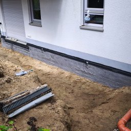 Lucjan Buduje - Wykop wokół ściany budynku z białą elewacją, ukazujący czarną folię kubełkową na fundamencie. W ziemi widoczne rury drenażowe, korytko odwadniające i narzędzia budowlane.
