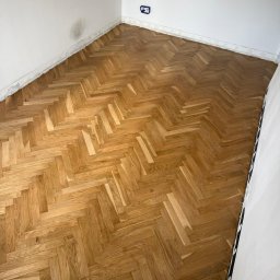 Bartosz Sztandera Atelier Parkietów - Odnowiony parkiet dębowy w jodełkę, z widocznym usłojeniem, w narożniku jasnego pokoju z białymi ścianami i gniazdkiem elektrycznym. Widoczne ślady prac wykończeniowych przy listwach.