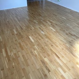 Bartosz Sztandera Atelier Parkietów - Jasna, błyszcząca drewniana podłoga z widocznym usłojeniem, ułożona w nieregularny wzór, w pustym pomieszczeniu ze świeżo pomalowanymi ścianami. W tle materiały budowlane.