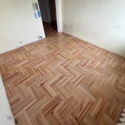 Bartosz Sztandera Atelier Parkietów - Nowo ułożony parkiet z jasnego drewna w jodełkę w pokoju z beżowymi ścianami, na których widać próbki farb, oraz otwartymi drzwiami.
