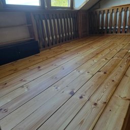 Bartosz Sztandera Atelier Parkietów - Jasne, drewniane deski podłogowe z widocznymi sękami w pomieszczeniu na poddaszu, z drewnianą balustradą i oknem ukazującym błękitne niebo oraz zielone drzewa.
