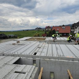 Grupa PHM - Trzech pracowników w kamizelkach odblaskowych i kaskach pracuje na stropie z płyt kanałowych, przygotowując zbrojenie do zalewania betonem. W tle zielone wzgórza i domy jednorodzinne...