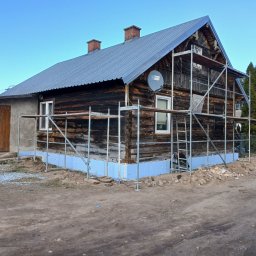 KS-DOM - Drewniany dom z metalowym dachem i dwoma kominami, częściowo ocieplany niebieskimi płytami izolacyjnymi u podstawy, otoczony rusztowaniem. Widoczne okna i drewniane drzwi.