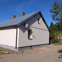 KS-DOM - Jasna elewacja domu jednorodzinnego z szarym dachem, brązowymi rynnami i ciemnym cokołem, widoczna antena satelitarna, w słoneczny dzień.