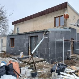 KS-DOM - Dom z czarnymi płytami izolacyjnymi na parterze i rusztowaniem, pracownik przygotowuje materiały. Górna część elewacji beżowa, dach blaszany. Zima, bałagan budowlany.