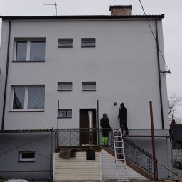 KS-DOM - Jasnoszara elewacja dwupiętrowego domu, dwóch pracowników na rusztowaniu finalizuje prace przy wejściu z ozdobną balustradą. Białe okna, ciemne rynny, pochmurne niebo.