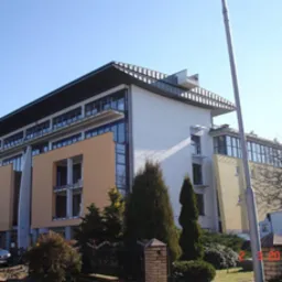 Budynek usługowy z elementami w kolorze żółtym i pomarańczowym, widoczny z zewnątrz, z przodu kilka krzewów, w tle błękitne niebo.