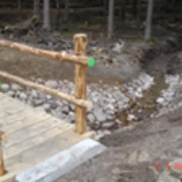 Drewniany mostek nad strumieniem w lesie, z barierami z surowych gałęzi, widoczne kamienie w korycie rzeki i leśne otoczenie.