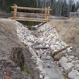 Mały mostek z drewnianą barierką nad strumieniem z brzegami umocnionymi kamieniami, widoczne elementy konstrukcji hydrotechnicznej.