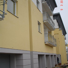 Fragment elewacji budynku wielorodzinnego w trakcie prac wykończeniowych, widoczne rusztowania i pracownik w tle. Ściany pomalowane na żółto, szare balkony z metalowymi balustradami.