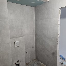 Paweł - Szara łazienka w trakcie wykończenia, z matowymi płytkami ściennymi i wzorzystą podłogą. Widoczna spłuczka podtynkowa, otwory instalacyjne i sufit z płyt GK.