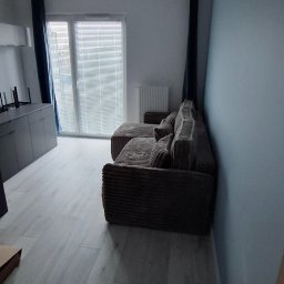 Paweł - Nowoczesne wnętrze z ryflowaną, brązową sofą narożną, jasną podłogą panelową i czarną lampą wiszącą w kształcie sześcianów. Ściana w odcieniu błękitu, okno z żaluzjami i granatowe zasłony.