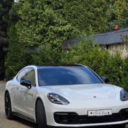 Artur Kubicki - Białe Porsche Panamera z czarnym dachem, ozdobione białymi wstążkami i tablicą 'Młoda Para', zaparkowane na brukowanym podjeździe wśród zieleni.