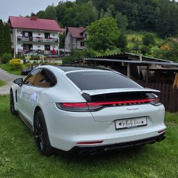Artur Kubicki - Białe Porsche Panamera z czarnymi felgami i czerwoną listwą LED, ozdobione wstążkami, z tablicą 'Młoda Para', zaparkowane na trawie przed domem.