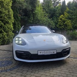 Artur Kubicki - Biała Porsche Panamera z tablicą 'Młoda Para' i włączonymi światłami, zaparkowana na brukowanym podjeździe, otoczona zielonymi drzewami i krzewami.