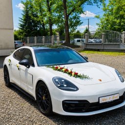 Artur Kubicki - Białe Porsche Panamera, udekorowane czerwono-żółtymi kwiatami na masce i lusterkach, z tabliczką 'Ania & Tomek', zaparkowane na żwirowym podjeździe w słoneczny dzień.