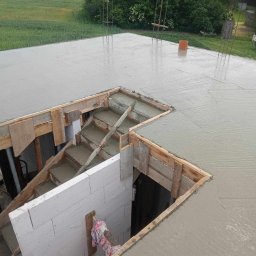 J&K Józefko - Świeżo wylana płyta stropowa z mokrego betonu, z widocznym otworem na schody, gdzie betonowe stopnie są uformowane w drewnianym szalunku. Obok ściana z białych bloczków, w tle zielony krajobraz.