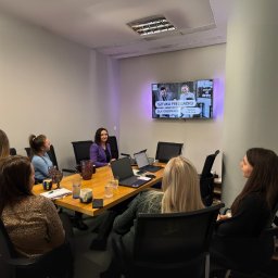 Hive Tax - W biurze w Warszawie, grupa kobiet przy drewnianym stole z laptopami ogląda na TV prezentację 'Sztuka Feedbacku dla Księgowych'. Kobieta w fioletowej marynarce patrzy na uczestniczki.