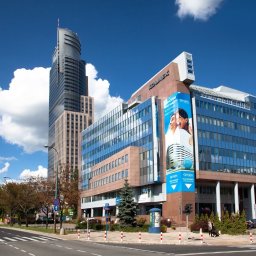 Hive Tax - Nowoczesny kompleks biurowy z niebieskimi elewacjami i reklamą 'Oxygen' na tle wysokiego wieżowca, przy ulicy z przejściem dla pieszych, w słoneczny dzień.