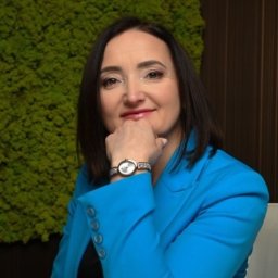 Hive Tax - Kobieta w turkusowym blazerze, z ręką na brodzie, uśmiecha się na tle zielonej ściany z mchu i drewnianego panelu.