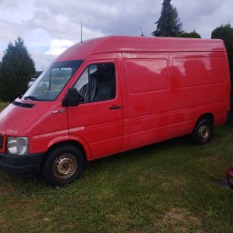 Paweł Kuźmiński, Przeprowadzki i Porządki- usługi transportowe - Czerwony bus dostawczy Volkswagen LT 35, zaparkowany na trawniku, z widocznymi zardzewiałymi felgami, pod zachmurzonym niebem i zielonymi drzewami w tle.