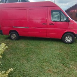 Paweł Kuźmiński, Przeprowadzki i Porządki- usługi transportowe - Czerwony furgon dostawczy Volkswagen LT 35 z wysokim dachem, zaparkowany na zielonym trawniku przed domem, pod zachmurzonym niebem.