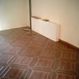 Ułożona brązowa podłoga z płytek w geometryczny wzór w pustym pokoju. Widoczne niedokończone krawędzie przy ścianach i listwa przypodłogowa. Biały grzejnik na ścianie.