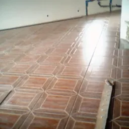 Układanie brązowych płytek podłogowych o geometrycznym wzorze w pomieszczeniu w Czarnkowie, widoczna listwa przypodłogowa i fragment kominka z kamienia.