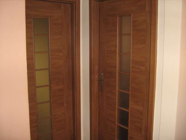 Dwie drewniane drzwi wewnętrzne z przeszkleniami w pionowych panelach, widoczne zawiasy i klamka, ściana w odcieniach bieli i różu.