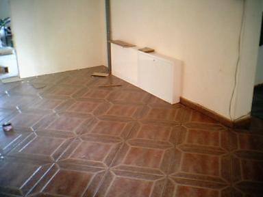 Ułożona brązowa podłoga z płytek w geometryczny wzór w pustym pokoju. Widoczne niedokończone krawędzie przy ścianach i listwa przypodłogowa. Biały grzejnik na ścianie.