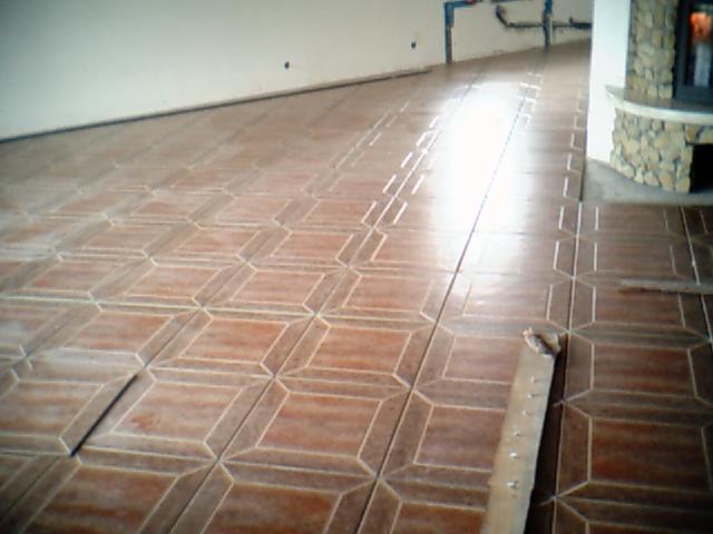 Układanie brązowych płytek podłogowych o geometrycznym wzorze w pomieszczeniu w Czarnkowie, widoczna listwa przypodłogowa i fragment kominka z kamienia.