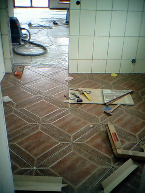 Układanie brązowej, wzorzystej terakoty w łazience, widoczne narzędzia glazurnicze i odkurzacz przemysłowy, płytki ułożone do połowy pomieszczenia.